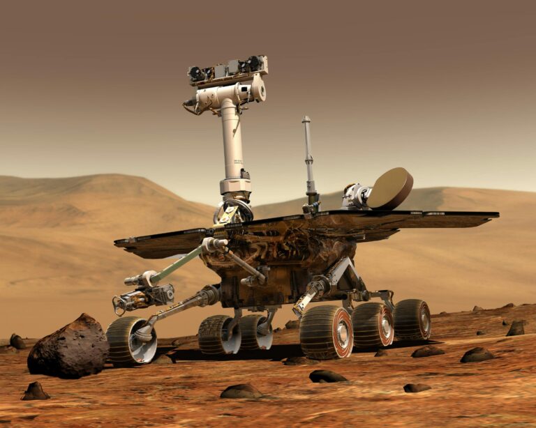 White and black NASA Mars rover with robotic arm parked on reddish rocky terrain under dusty pink sky